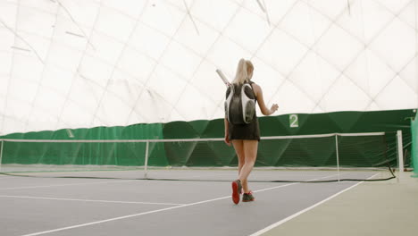 TRACKING-Young-Caucasian-female-tennis-player-entering-the-court,-rackets-in-a-backpack.-4K-UHD-RAW-graded-footage